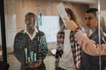 A multi-ethnic group of business professionals collaboratively analyze and strategize using diagrams and sticky notes on a transparent wall in modern office setting.