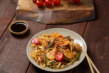 udon shrimp in pad thai sauce on a wooden table next to cherry tomatoes, soy sauce and chopsticks
