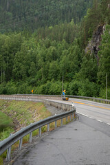 mountain road in Norway whose lanes are separated by a solid yellow line and which runs next to mountains with green coniferous trees.