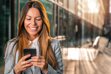 Portrait of beautiful business woman standing in front of her office building and smiling while reading text on her mobile phone.