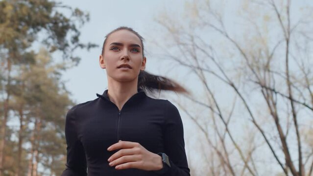 Young caucasian female jogger in a black sportswear running with a confident face on an alley in an autumn city park close up, bottom view. Concept of modern outdoor sports training in a casual urban