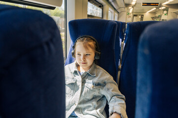 A little traveler is sitting on a blue chair on a train. Little girl in headphones looking out the...