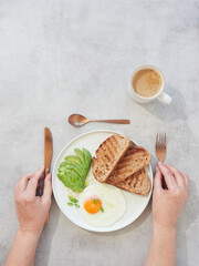 Hands holding cutlery, knife and fork, next to plate with toast, egg and avocado. Healthy breakfast with coffee.