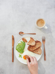 Chunk of toast in hand dipping the yolk of an egg. Healthy breakfast plate with avocado on the table with a cup of coffee.