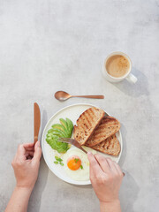 Hands holding fork over plate to eat avocado, egg and toast. Healthy breakfast on table with cup of coffee.