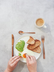 hands with pieces of bread dipping in the yolk of an egg. Healthy breakfast plate with avocado on the table with a cup of coffee.