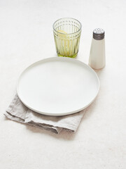Table setting, empty white plate on napkin, salt shaker and green clear glass with lemon. High angle view on bright background.