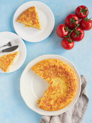 Typical homemade Spanish potato omelette, triangular portions served on plates. Fresh red tomatoes on blue background.