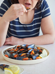 woman opening her mouth to eat a mussel in its shell in front of a large platter with cooked mussels and fresh lemon slices