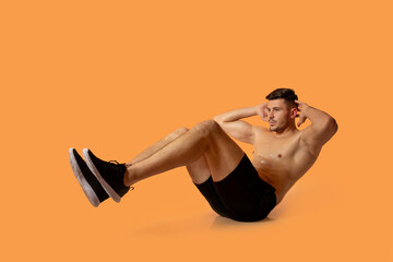 A fit young man is doing bicycle crunches against a solid orange background in a studio, showcasing...