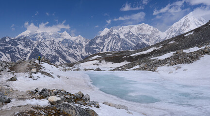 Fototapeta premium A hiker on a trail to the Tilicho Lake, Annapurna Conservation area, Nepal. A small lake covered by ice. A trail covered by snow and ice. Frozen lake. Mountain range in the background.