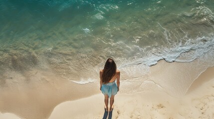Aerial view of a white sandy beach with white waves rolling onto shore and crystal clear blue water and a girl standing on the beach casting a long shadow,