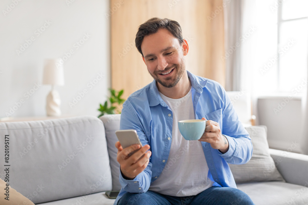 Poster a man is comfortably seated on a couch, holding a cup of coffee in one hand and using a smart phone 
