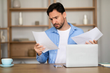 A man is sitting at a desk at home office, looking with a perplexed expression at some documents in...