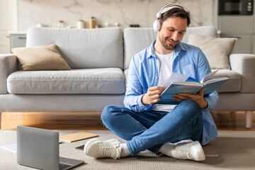 A man is seated on the floor, engrossed in reading a book while simultaneously listening to music through headphones. He appears focused and relaxed in his dual activities.