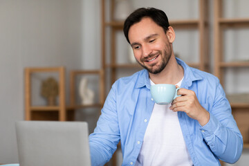A man is seated at a desk, using a laptop while sipping on a cup of coffee. He appears focused on...