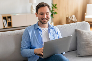 A man is sitting on a couch while using a laptop. He appears focused on the screen, typing and clicking. The room is well-lit, with a coffee table in front of him.