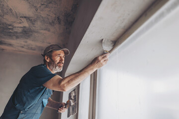 Construction worker wearing worker overall with wall plastering tools renovating apartment house. Plasterer renovating indoor walls and ceilings with float and plaster. Construction finishing works.