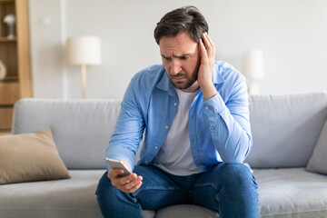 A man appears troubled or exasperated, sitting with a slouched posture on a grey couch. He cradles his head in one hand, with a smartphone held in the other. His expression conveys distress or concern