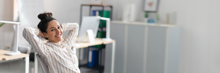 Happy relaxed lady with hands behind her head enjoying rest from work in office interior, sitting at workplace and smiling at camera. Successful business and great deal, copy space