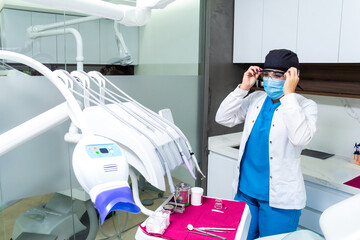 Female latin hispanic dentist using dentistry uniform and protective safety clothing, standing next to a dental chair. Clinic or hospital concept