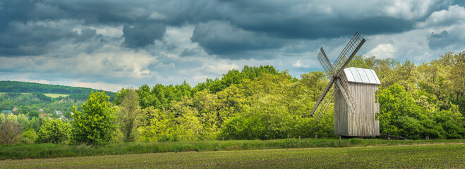 wide angle panoramic view to farmland landscape with old wooden windmill and dark clouds in spring...