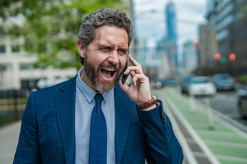 Anger Businessman with phone outdoor. Angry Business talk. Anger Man walk down street talking on...