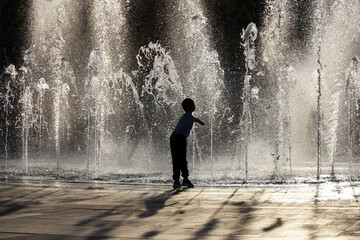 Silhouette of kid boy plays with water jets of fountain. Child enjoying the summer, hot weather in...