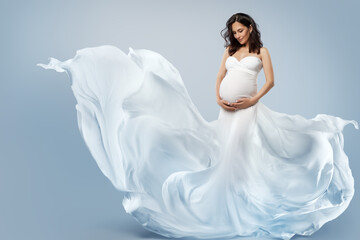 Pregnant Woman in White Fantasy Dress flying on Wind. Beautiful Mother looking at Belly in Silk...