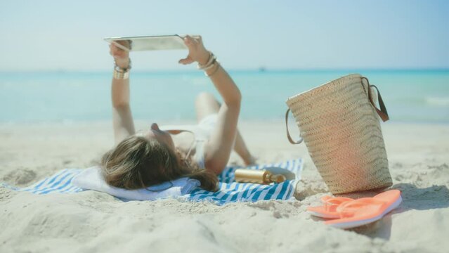 relaxed elegant 40 years old woman on the seacoast with straw bag and striped towel digital tablet blank screen.