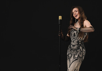 Beautiful young woman in stylish dress with microphone singing on black background
