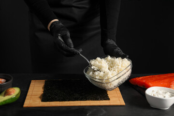 Chef in gloves taking cooked rice for sushi with spoon at dark table, closeup
