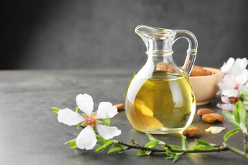 Almond oil in jug, branch with flowers and nuts on grey textured table, closeup. Space for text