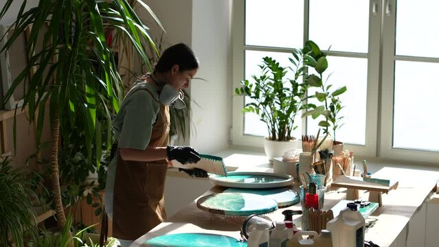 Woman artist examining finished acrylic sea painting on wooden round canvas covered with epoxy resin. Painter works in art studio with indoor plants. Home art studio, small business, hobby, liquid art