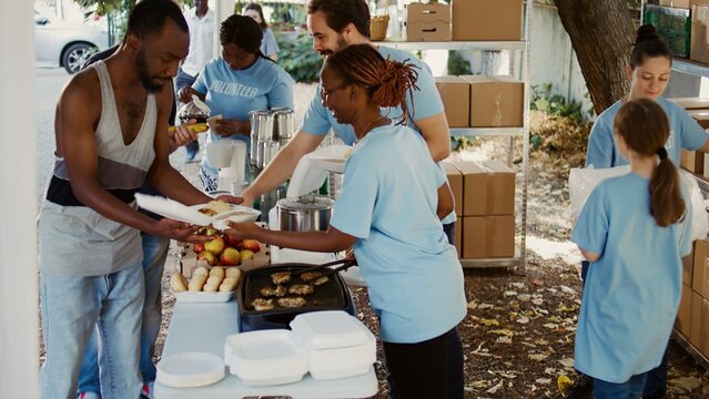 At local center, group of volunteers arranging donation boxes and preparing hot meals for individuals facing hunger and poverty. Youthful charity workers handing out free food to the needy. Handheld.