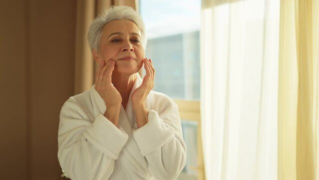 Mature adult old woman applying nourishing anti age face cream doing daily beauty routine. Senior woman touching face skin wrinkles gets facial treatment. Skin care aging beauty skincare cosmetics