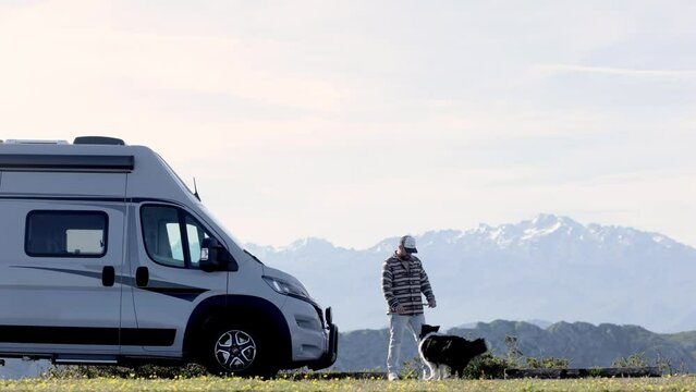 Man in jacket playing with his dog nearby a white van against mountains, contemplate thoughts and find calm peace. Camping trip in great outdoors. Van lifestyle. Family vacation travel