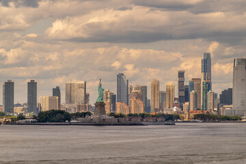 Obraz premium New York, NY, USA - August 1, 2023: Statue of Liberty and its green island under blue cloudscape. Skyline of Jersey City with wall of tall buildings