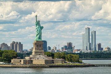 New York, NY, USA - August 1, 2023: Jouned Squared tower of Jersey city is seen to the right of Statue of Liberty on its island under blue cloudscape. Plenty of visitors and more building in back