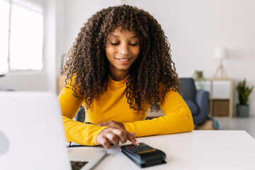 Young african american woman using a calculator while working on laptop at home. Business and education concept.