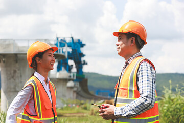 Senior construction male  engineer training a young new female  engineer and using a digital tablet with a hold walkie-talkie in his hand at the infrastructure of Sky bridge