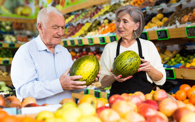 Woman supermarket salesman helping elderly man to choose watermelon and other fruits