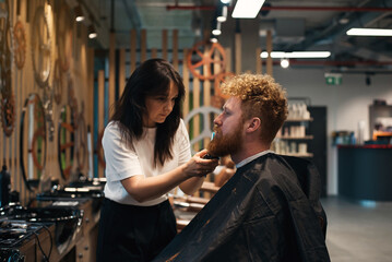 Red-haired bearded man sitting in barbershop and female barber making hairdo to guest