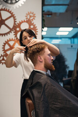 Red-haired bearded man sitting in barbershop and female barber making hairdo to guest