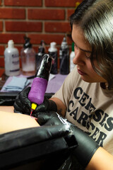 Young adult female tattoo artist using professional tattoo machine to create tattoo in client's...