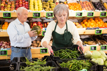 Obraz premium European mature woman greengrocer worker in apron standing in salesroom and setting out goods on shelves. Old man with melon in hands standing behind.