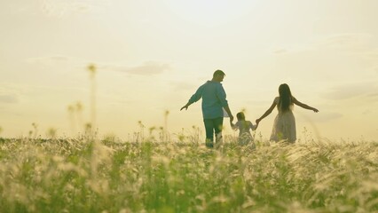 Happy family running through grass in meadow in summer at sunset, rear view, slow motion. Father,...