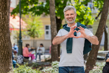 Fototapeta premium I love you. Caucasian middle-aged man makes symbol of love showing heart sign to camera, express sincere romantic positive feelings. Charity, gratitude, donation. Mature guy on urban city street park.