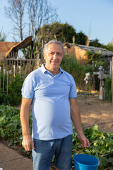 Portrait of positive senior man standing in backyard garden in village, holding bucket, looking at camera and smiling.