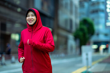 Glad smiling young asian muslim female in hijab with fitness tracker running on building background...